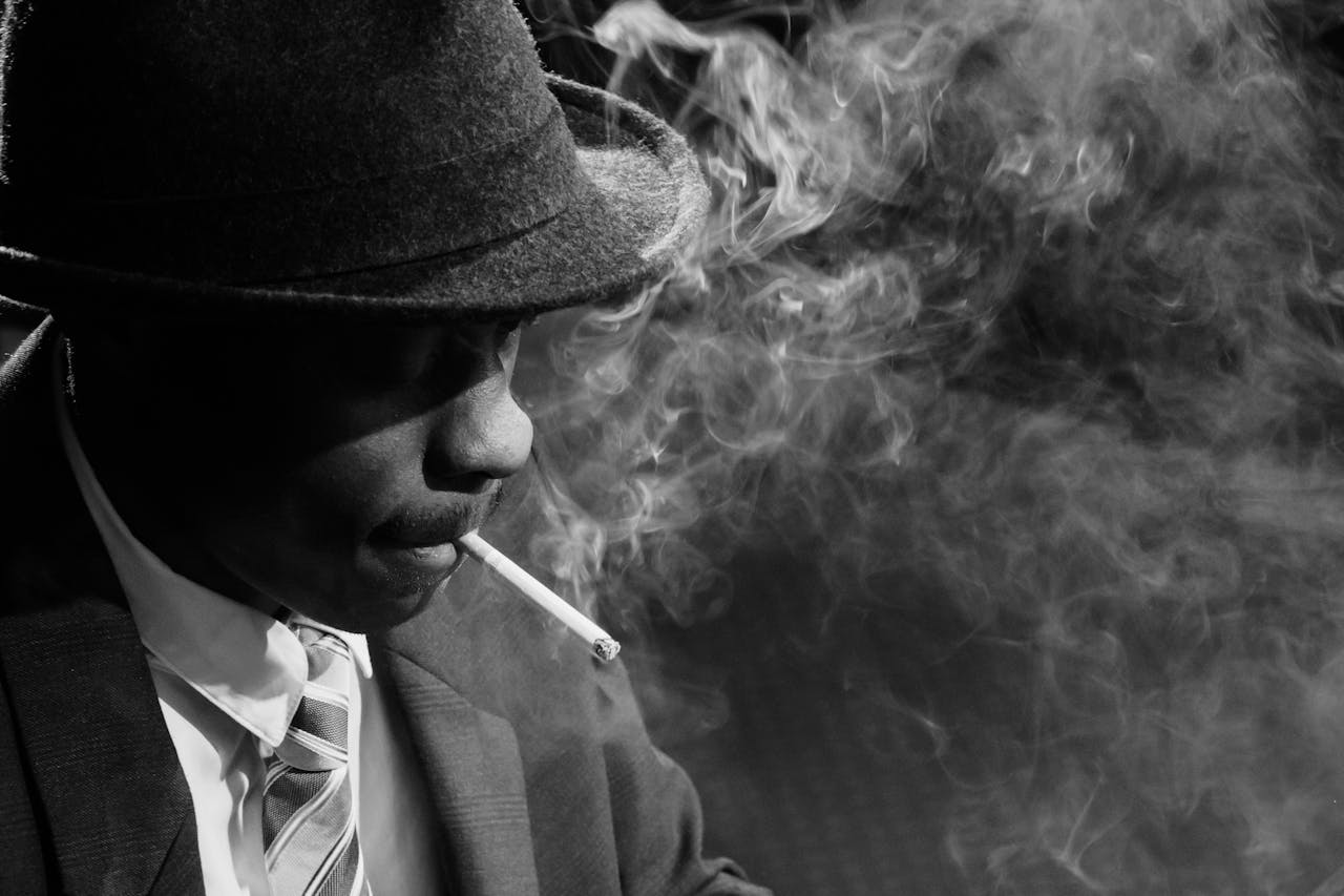 A black man in 1950s noir style with a fedora hat, smoking a cigarette amidst swirling smoke.